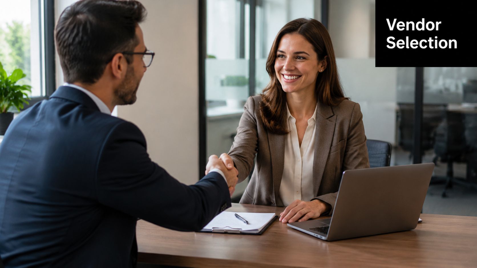 A professional man and woman shaking hands over a wooden office desk after a business agreement.